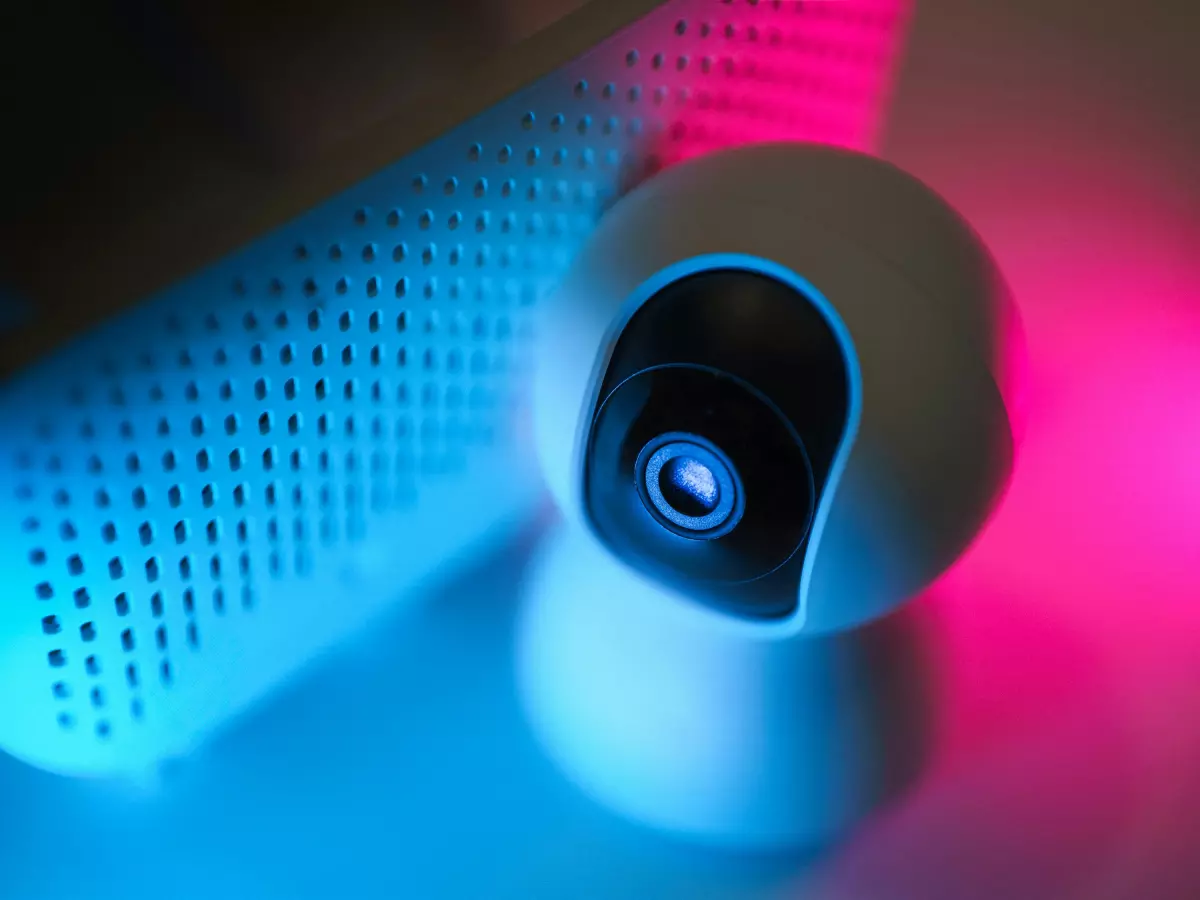 A close-up of a white security camera with a black lens, lit by blue and pink neon lights.